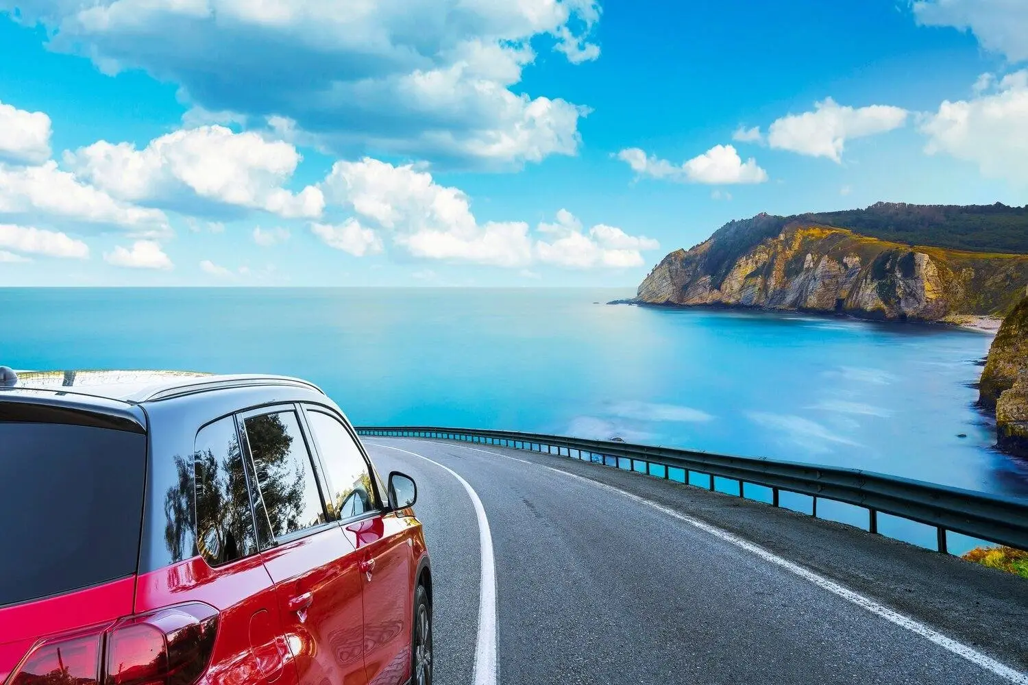 Red car driving on a coastal road overlooking the blue ocean and rocky cliffs under a bright sky with clouds